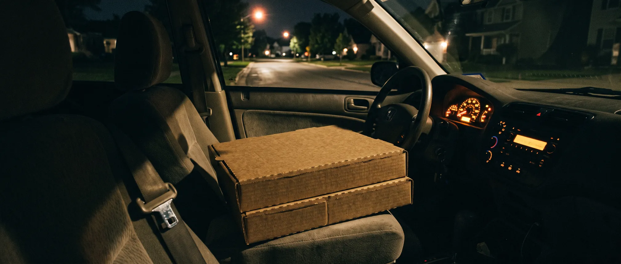 Two plain brown pizza boxes stacked on a car passenger seat at night, lit by the dashboard and the smear of oncoming headlights.